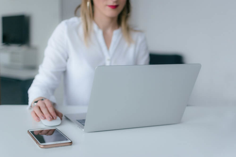 PearlConvert markedsføring Kvinne i hvit skjorte og rød leppestift sitter med sin datamaskin og styrer den med datamus, med hennes iPhone liggende ved siden av.